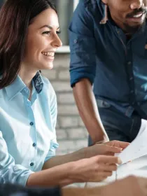 A group of four people in a modern office setting, smiling and discussing while looking at a computer screen. One person is holding a piece of paper.