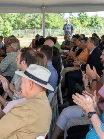 A large group of people seated under a tent, attentively watching an event and applauding. The audience includes men and women of various ages, some wearing hats. The background shows greenery and a fence.