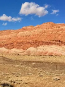 Red rock formations under a clear blue sky with scattered clouds, set in a dry, grassy landscape.