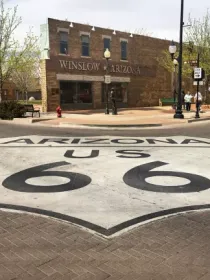 A street view of Winslow, Arizona, featuring a large Route 66 emblem painted on the road. The historic brick buildings and parked vehicles are visible under a partly cloudy sky.