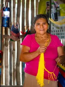 A woman in a pink shirt stands in a rustic room, weaving a colorful hammock. She holds strands of yellow yarn, surrounded by wooden walls and a hanging lantern.
