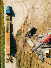 Aerial view of a construction site with a blue truck, a yellow trailer, and a red crane. The site is adjacent to a green field.