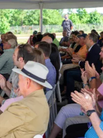 A group of people seated under a tent at an outdoor event, clapping. They are sitting on white folding chairs, with trees visible in the background.