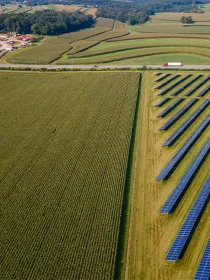 Aerial view of a landscape with solar panels aligned on a field next to a large cornfield. Rolling hills and a road with vehicles are visible in the background.