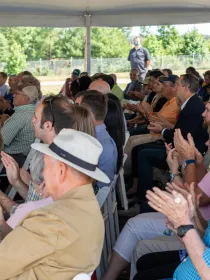 A diverse audience sitting under a tent applauding during an outdoor event. The crowd is seated in rows of white chairs, with trees and a fence visible in the background.