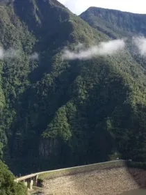 A lush green mountain landscape with a dam at the base, surrounded by dense forest and a few clouds drifting across the sky.