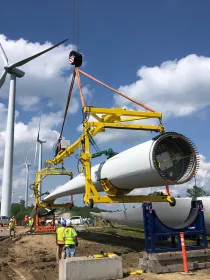 Workers in safety vests assemble a large wind turbine blade using a crane at a construction site with multiple wind turbines in the background under a blue sky with clouds.