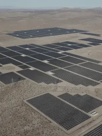 Aerial view of a large solar farm in a desert landscape. The solar panels are arranged in a grid pattern, covering a vast area of the barren land. Sparse vegetation and distant mountains are visible in the background under a clear sky.