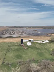 Aerial view of a large solar farm with rows of solar panels on a vast field. In the foreground, there is a small farm with a barn and silo surrounded by green fields and trees. The sky is partly cloudy.