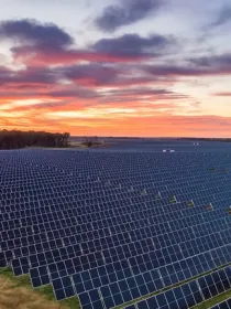 A large field of solar panels under a vibrant sunset sky. The panels are arranged in neat rows, stretching across the landscape. The sky is filled with orange, pink, and purple hues, creating a striking contrast with the dark panels.