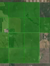 Aerial view of farmland with green fields divided into rectangular plots. Some plots show distinct patterns of cultivation, while others appear more uniform. Surrounding areas include patches of brown and gray land, possibly indicating uncultivated or harvested areas.