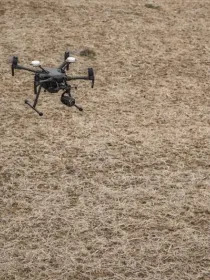 Two construction workers in safety gear operate a drone over a dry, barren field.