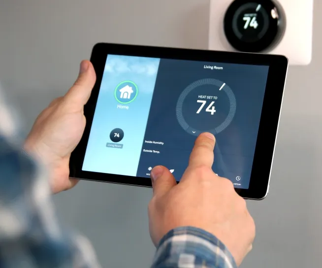 Person adjusting a smart thermostat using a tablet. The screen shows a temperature setting of 74 degrees for the living room. A wall-mounted thermostat displays the same temperature.