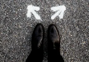 A person wearing black shoes stands on a gravel road in front of two white arrows painted on the ground, pointing in different directions, symbolizing a choice or decision.