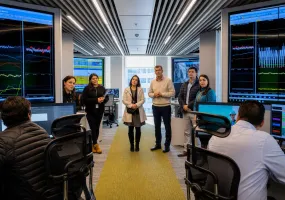 A group of professionals standing in a modern control center filled with monitors displaying data graphs.