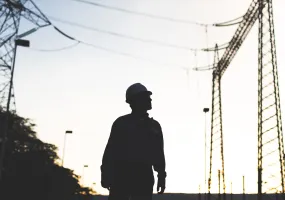 Silhouette of an electrical worker wearing a hard hat standing in front of high voltage transmission towers at sunset.