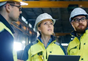 Three construction workers wearing safety helmets and high-visibility jackets stand inside a large industrial facility. One holds a laptop, and they appear to be discussing or inspecting something above.