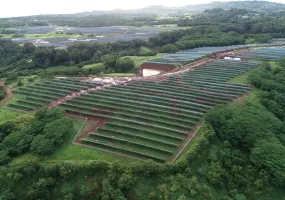 Aerial view of a large solar farm with rows of solar panels on a green landscape surrounded by trees and hills.