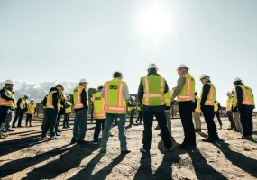 Group of construction workers in high-visibility vests and hard hats standing in a circle on a sunny day, with mountains in the background.