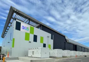 A modern industrial building with a geometric design and the letters 'aes' on the facade. The building features a mix of white, green, and dark panels, with several electrical units in front. The sky is partly cloudy.