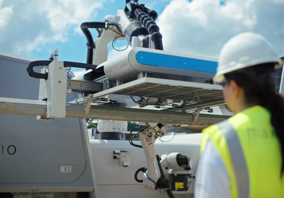A person wearing a hard hat and high-visibility vest observes a robotic machine installing a solar panel under a partly cloudy sky.