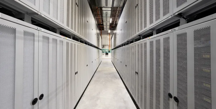 A narrow hallway in a data center with rows of server racks on both sides. The floor is concrete, and the ceiling has visible cables and lights.