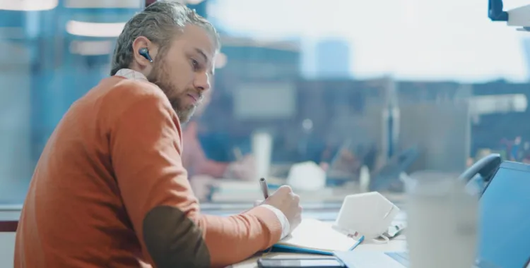 Man with earbuds writing in a notebook at a desk with a laptop and smartphone.