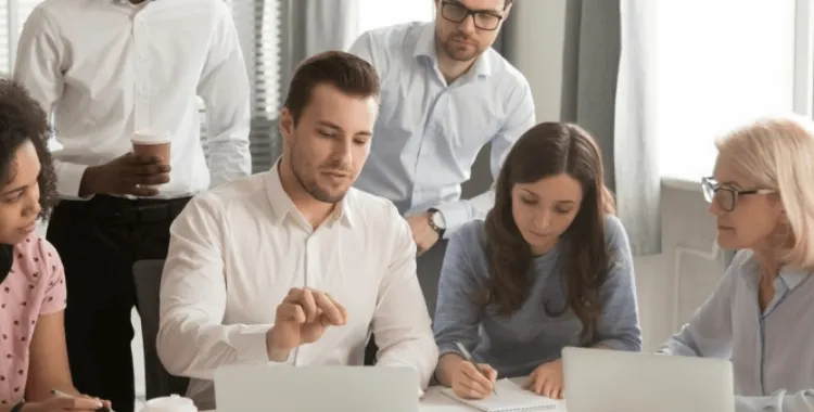 A group of six people in a meeting room, sitting and standing around a table. One person is explaining something while pointing at a laptop screen. Others are listening attentively, with one taking notes. The atmosphere appears collaborative and focused.