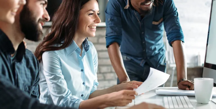 A diverse group of four young professionals collaborating in a modern office, smiling and looking at a computer screen.