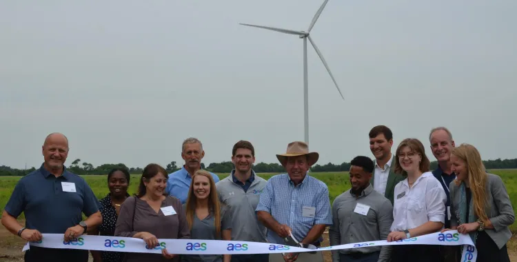 A group of people standing in front of a wind turbine, holding a ribbon labeled 'aes' during a ribbon-cutting ceremony in a field.