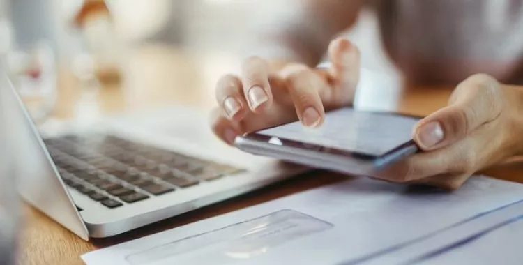 A person using a smartphone with one hand while another hand rests on a laptop keyboard. Papers are scattered on the desk, and the background is softly blurred.