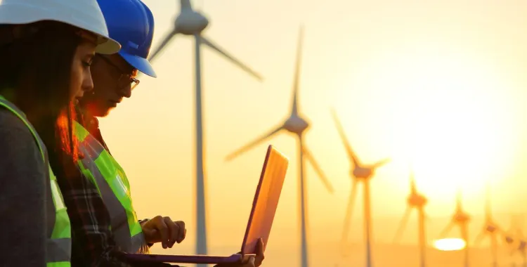 Two workers wearing safety helmets and reflective vests are using a laptop in front of a field of wind turbines during sunset. The sky is orange, and the sun is low on the horizon.