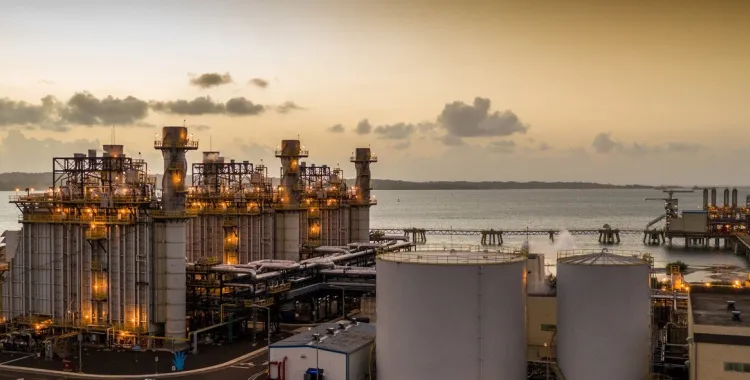 Industrial facility by the sea at sunset with large towers, pipes, and storage tanks, illuminated by lights.