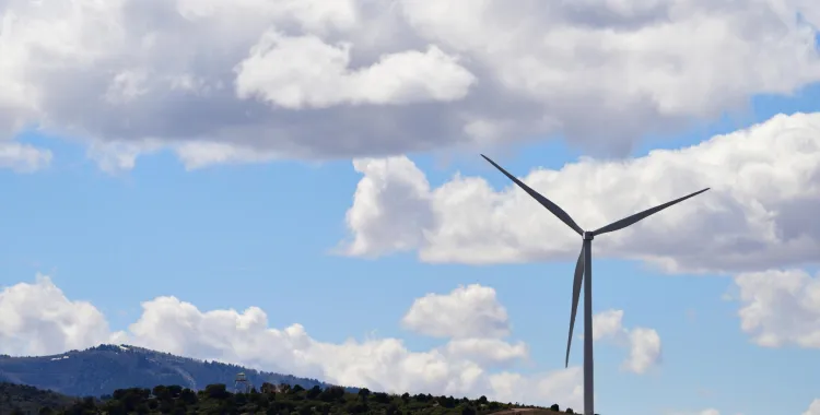 A wind turbine stands on a hill covered with green vegetation. The sky is bright blue with scattered fluffy white clouds. A mountain range is visible in the background.