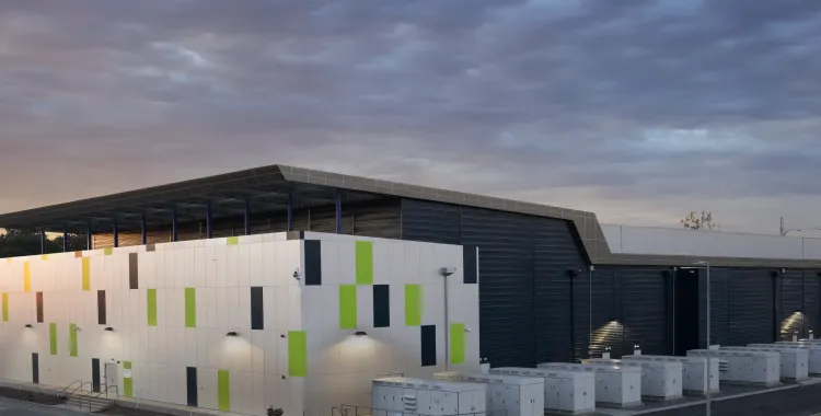 Modern industrial building with a geometric facade featuring green and gray accents, surrounded by rows of white utility cabinets, photographed at sunset under a cloudy sky.