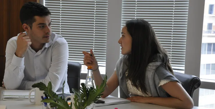 Two people sit at a table in a modern office, engaged in conversation. The woman gestures while speaking, and the man listens attentively. Office supplies, a potted plant, and name tags are visible on the table, with window blinds in the background.