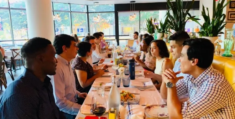 A group of people sit around a long table in a bright, modern restaurant, enjoying food and drinks while engaging in conversation. The table is set with plates, glasses, and flowers, and large windows let in natural light.