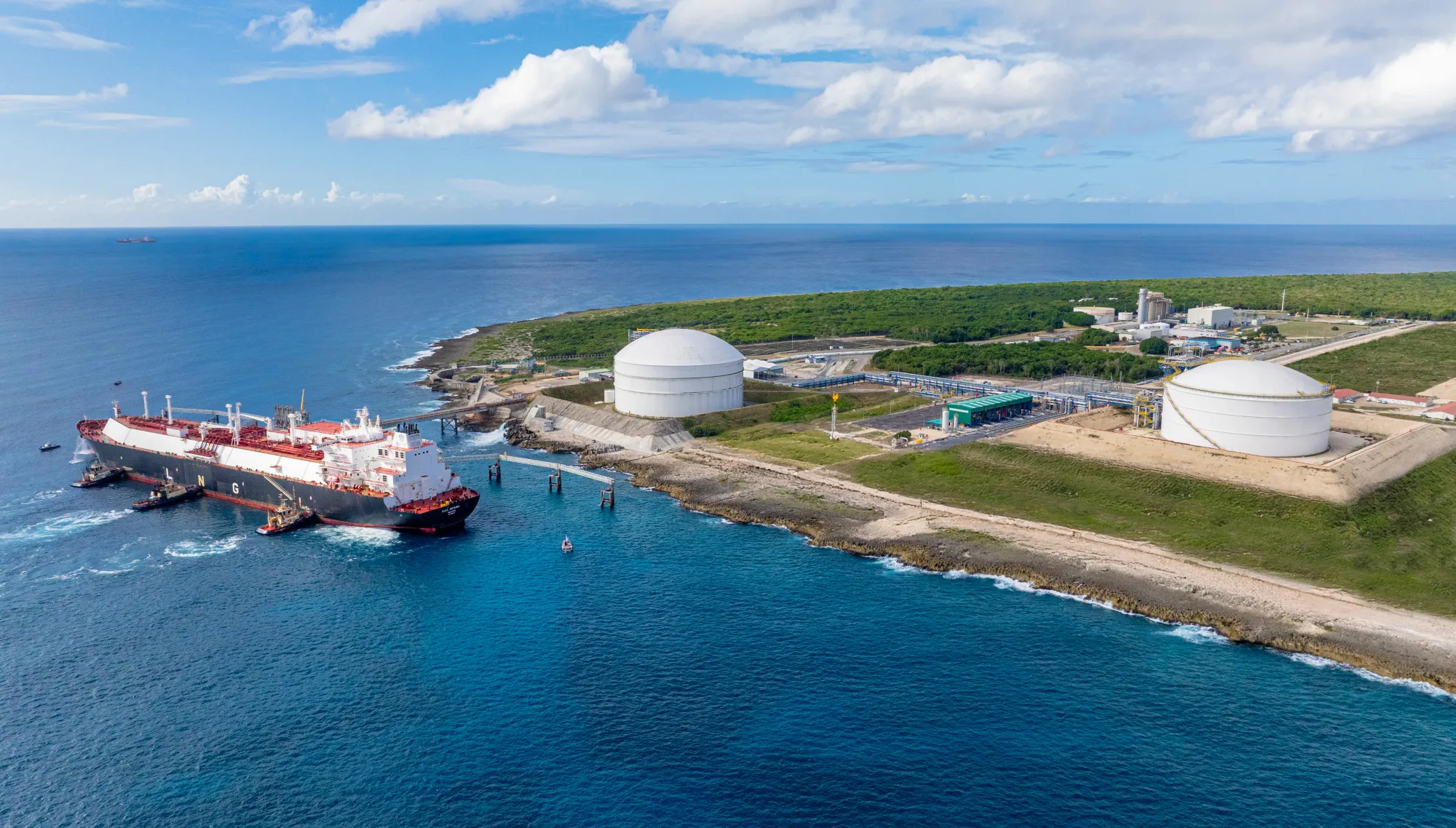 Aerial view of a large tanker ship docked at a coastal industrial facility with two large storage tanks, surrounded by greenery and ocean under a partly cloudy sky.