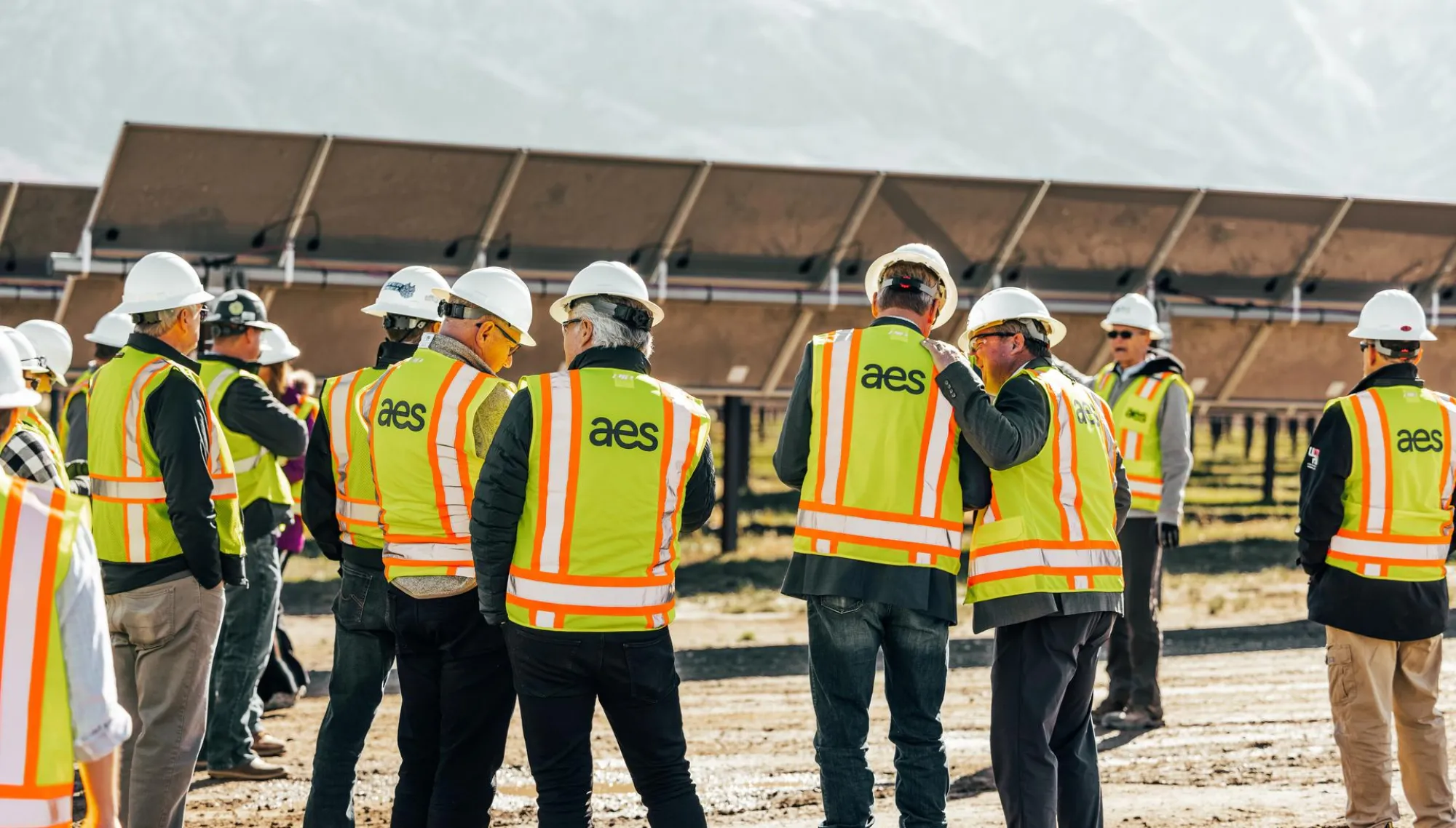 A group of people wearing AES-branded yellow safety vests and white hard hats stand on a dirt path in front of large solar panels. The background features a mountainous landscape.