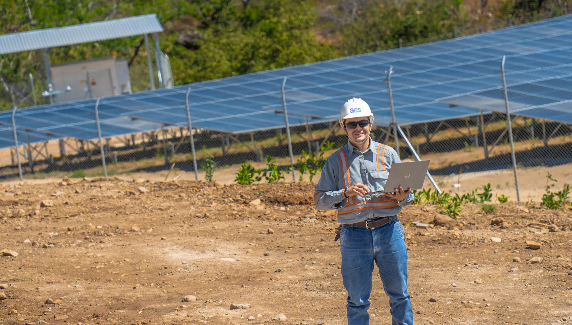 A person wearing a hard hat and safety vest stands on a dirt path holding a laptop. Behind them, a large solar panel array is visible, enclosed by a chain-link fence. Trees and greenery can be seen in the background.