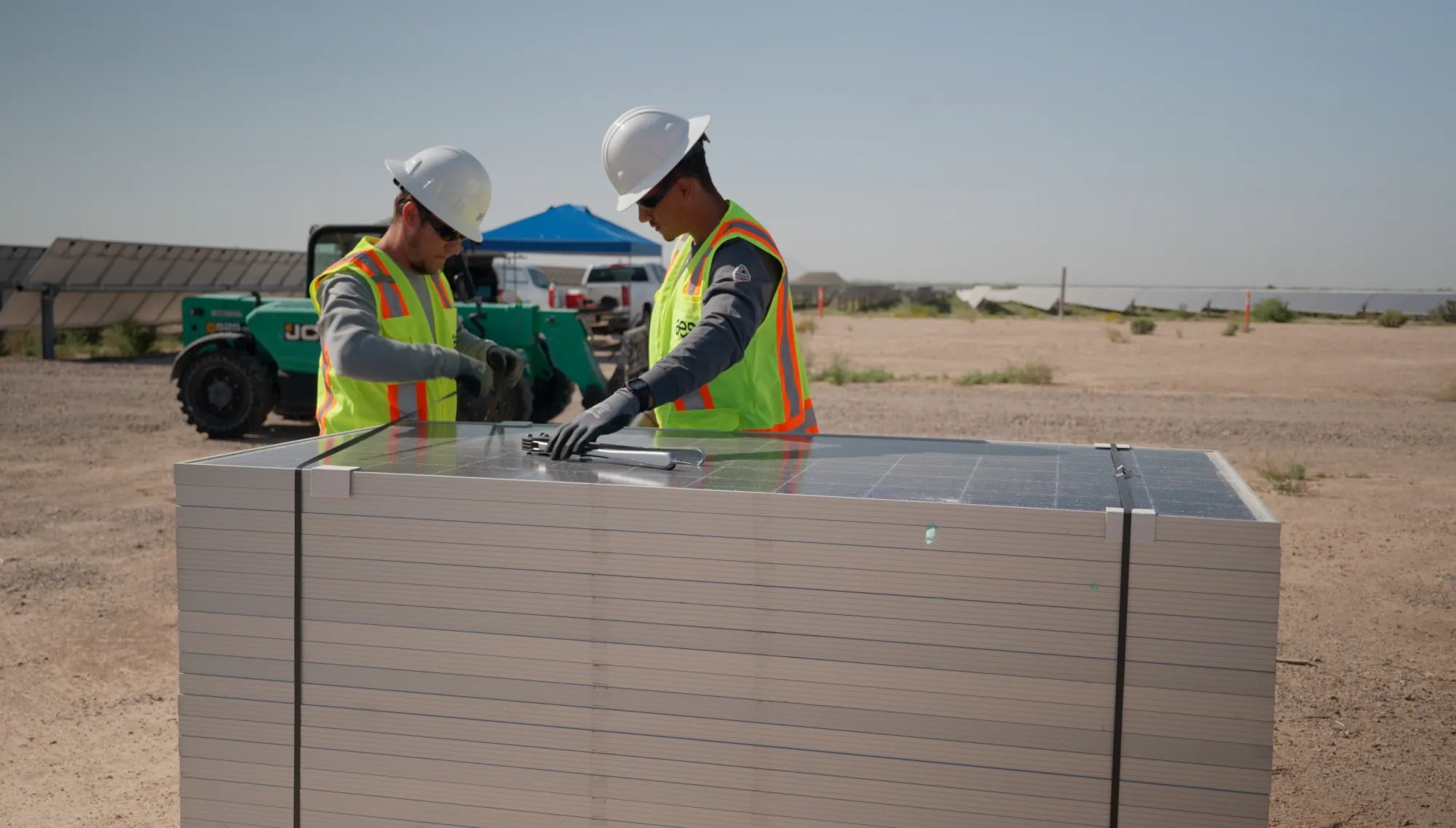 Two workers wearing safety vests and helmets are inspecting a stack of solar panels at a construction site. They are in a desert-like area with solar panels and machinery in the background.