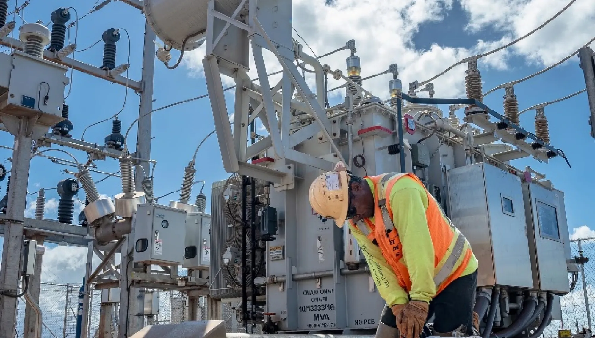 A worker in a safety vest and helmet kneels in front of electrical equipment at a power station under a partly cloudy sky.