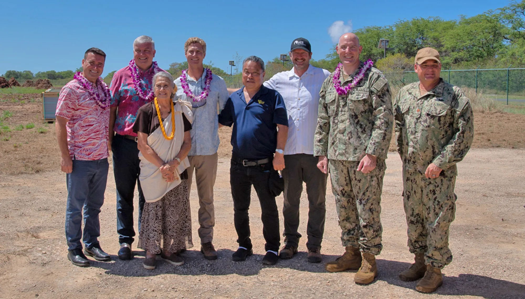 A group of eight people standing outdoors on a sunny day, some wearing leis and military uniforms, smiling at the camera with a clear sky and trees in the background.