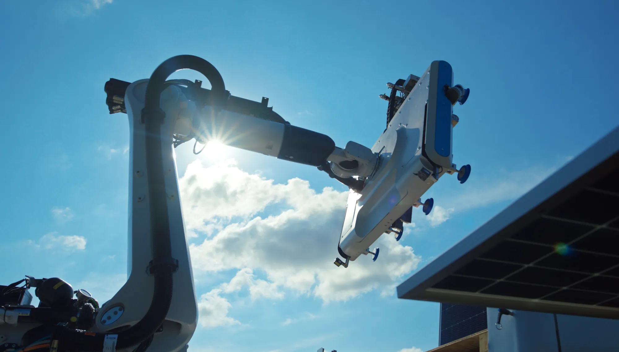 A robotic arm with a suction device operates under a bright blue sky with the sun shining through, highlighting its mechanical structure.