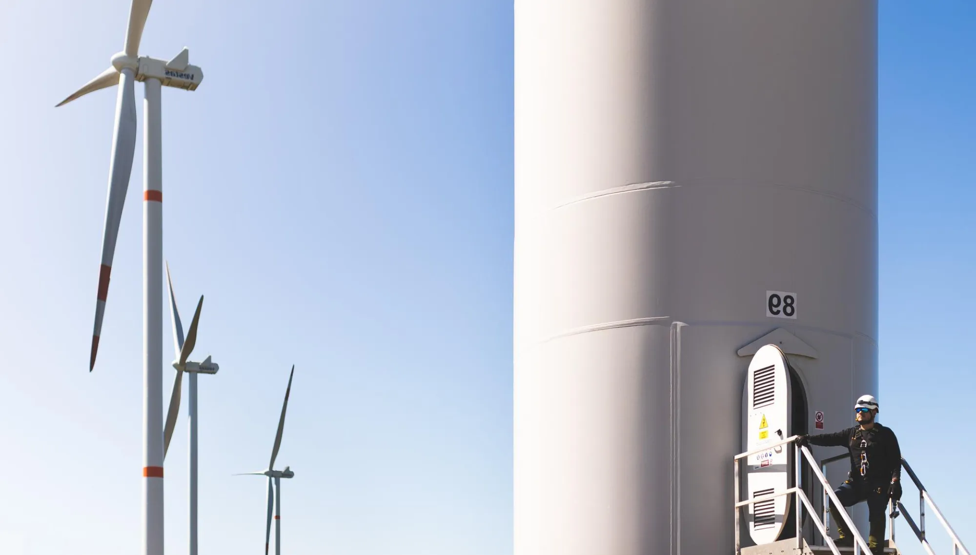 A worker in safety gear stands on a platform at the base of a large wind turbine. Several wind turbines are visible in the background against a clear blue sky.