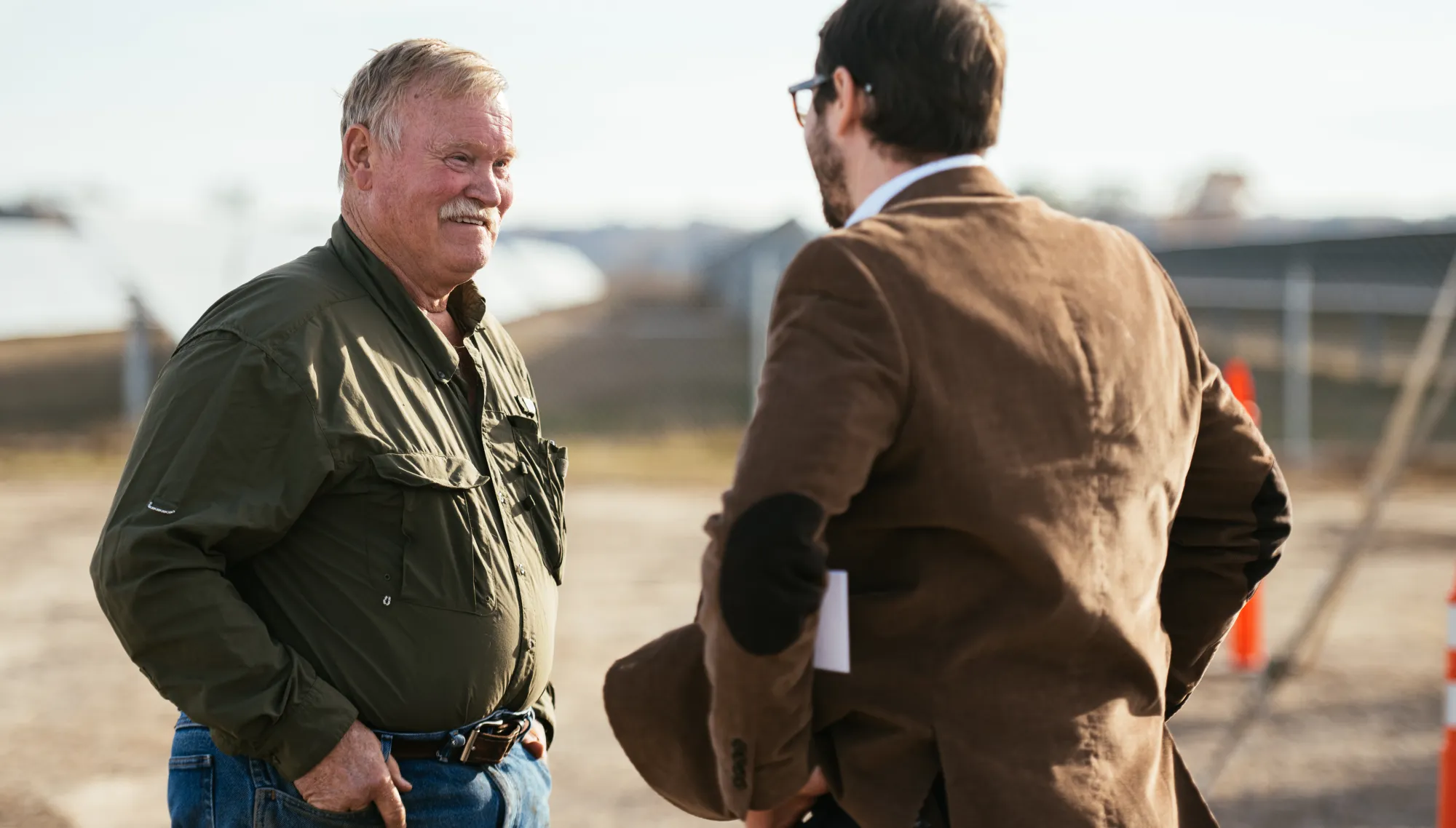 Two men are engaged in conversation outdoors. One man, wearing an olive green shirt and jeans, faces the camera. The other man, in a brown jacket with elbow patches, has his back to the camera. They are standing near a fenced area with blurred structures in the background.