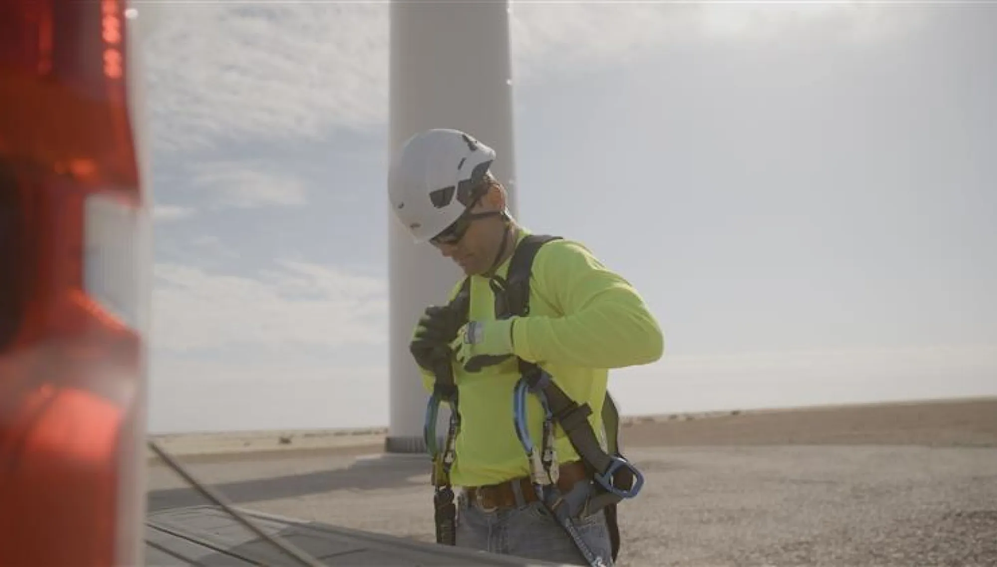 A person wearing a bright yellow safety jacket and helmet adjusts their harness near a wind turbine. The scene is outdoors with a clear sky.