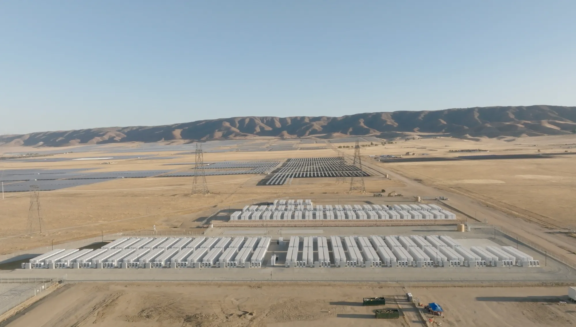 Aerial view of a large solar farm with rows of solar panels and battery storage units in a dry, open landscape. Power lines and distant hills are visible in the background under a clear blue sky.