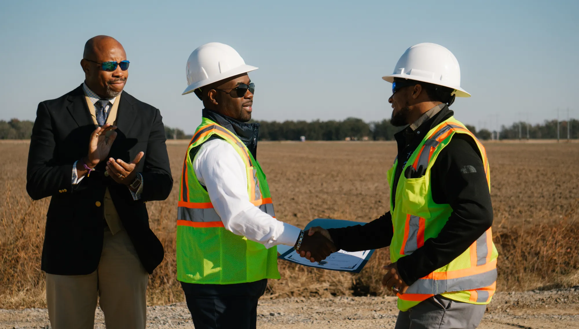 Three men at a construction site. Two men wearing hard hats and reflective vests shake hands, while a third man in a suit claps. The background shows an open field under a clear sky.