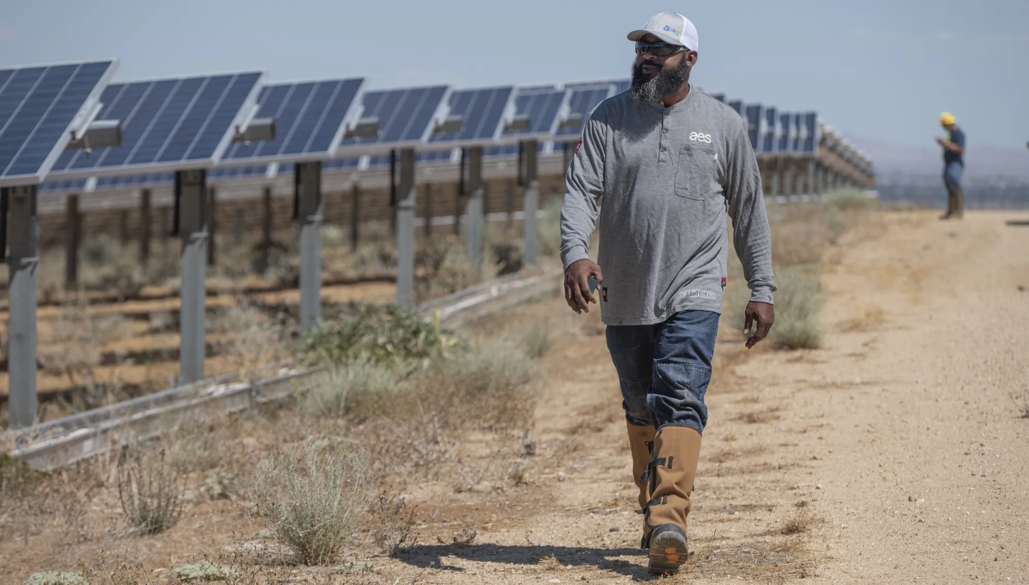A man in a gray shirt and brown boots walks along a dirt path beside a row of solar panels in a desert landscape. Another person is visible in the background near the panels.